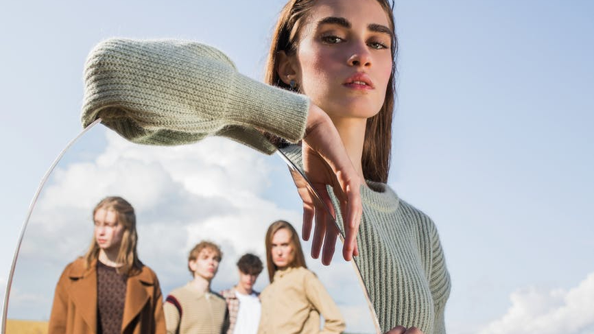 young woman standing in field with oval mirror reflecting group of young people By Halifax Tailor
